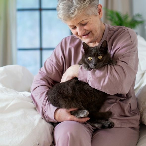 Ältere Frau in lila Nachtkleidung hält eine graue Katze liebevoll in ihren Armen.