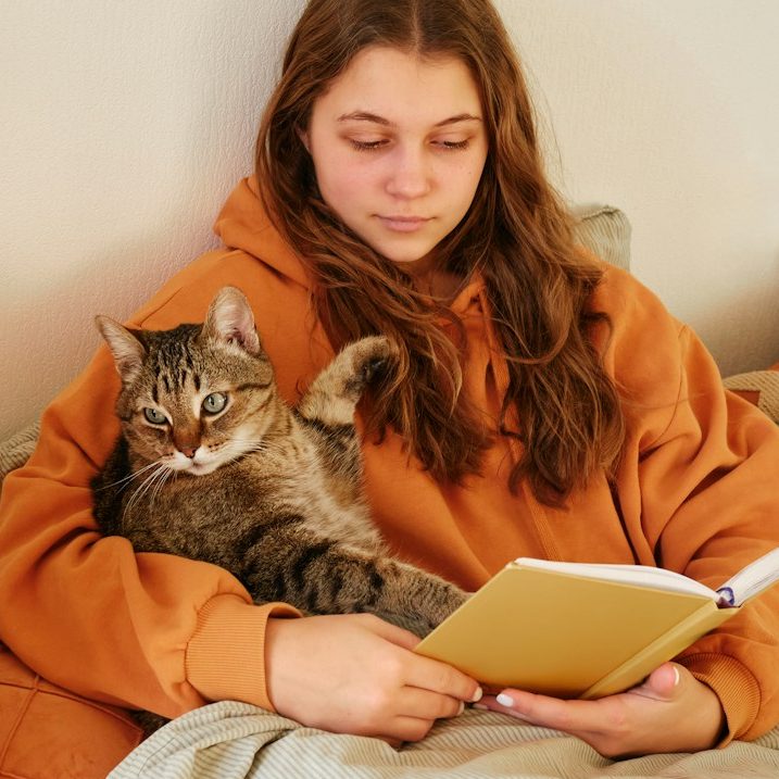 Mädchen in orangeem Hoodie liest ein Buch, eine Katze sitzt auf ihrem Schoß.